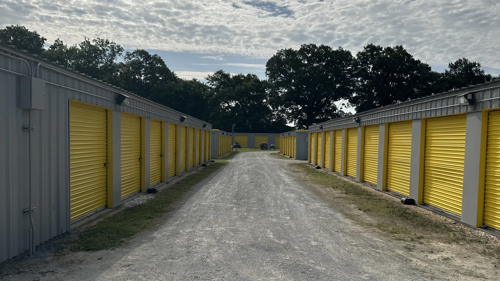 Rows of yellow roll-up unit doors lining both sides of a gravel drive aisle, with trees in the background